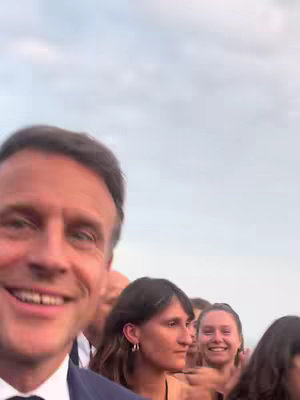 A selfie-style shot features Emmanuel Macron in the foreground, smiling directly at the camera. Behind him are a crowd of people, some smiling and looking in various directions. The background appears to be an overcast sky, creating a natural and informal setting.
