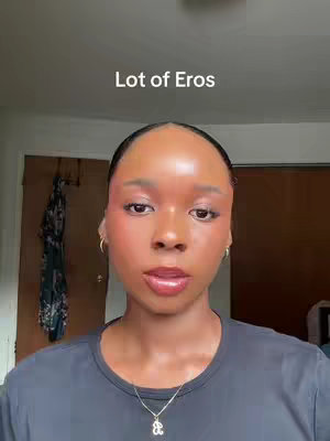 A close-up shot features a dark-skinned woman with sleek, black hair pulled back. Her skin glows with a subtle sheen, enhanced by blush and a glossy lip. She wears a black t-shirt and a delicate gold necklace. Behind her, out-of-focus doors and a patterned fabric hint at a domestic setting. The overlaid text 'Lot of Eros' is prominent at the top.