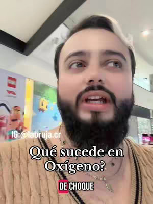 A medium shot of a light-skinned man with dark hair and a full beard. He is looking upwards and to his left, mouth slightly open as if he is speaking. The man is wearing a light-colored sweater with an indistinct background of a shopping mall or retail space. Text overlays in Spanish ask questions and provide de choque