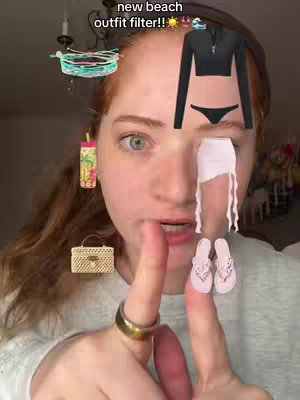 A close-up shot of a fair-skinned person with bright red hair and green eyes, suggesting a selfie style. Text overlayed says "new beach outfit filter!!". The person is looking toward the camera. Onscreen, a variety of beach-themed accessories hover near their face, including a straw bag, earrings, bracelets, and a range of outfit options.