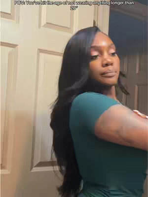 A woman stands in front of a bathroom door, angled slightly towards the camera. She has long, wavy black hair and is wearing a dark green top. She appears to be looking down, and has one arm resting on her chest. The background is a lightly colored door and a section of wall, neutral tones. Above her head is a white box saying 'POV: You've hit the age of not wearing anything longer than 20".