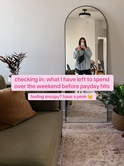 A selfie taken in front of a large, arched mirror. The mirror reflects a person holding a phone, likely taking the photo. The room has a neutral color palette, with a green sofa, a rug, and a plant visible.