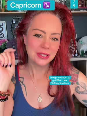 A woman with red hair is positioned slightly off-center, looking directly at the camera and speaking. She wears a dark blue tank top, and several visible tattoos are on her arm. She has bold eye makeup and red lipstick. A light blue banner at the top of the frame reads \"Capricorn\" in white font, accompanied by the Capricorn astrological symbol. Additional text overlaid on the image includes a banner stating \"Things are about to get REAL clear Shifting timelines\"