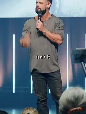A man wearing a gray t-shirt and black jeans stands on a stage. The man is holding a microphone, and the background consists of vertical white lights.