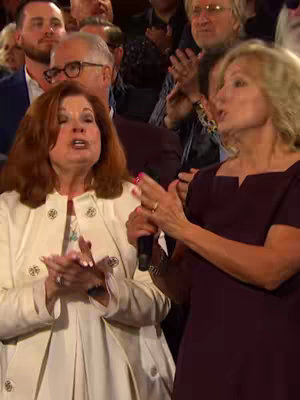 The image shows two women standing close together, presumably in an auditorium or large event space, given the crowd visible in the background. Both women are facing forwards and appear to be singing or speaking, with microphones in front of them. The woman on the left has red hair and is wearing a white coat over a patterned top. The woman on the right has blonde hair and a dark purple dress. The audience in the background is blurred, with some individuals clapping.