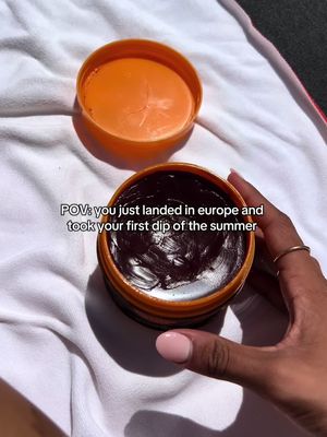 A close-up shot shows an open orange tub with a dark, creamy substance inside, sitting on a white towel. A hand with pink nail polish and a gold ring is holding the tub, and the background includes a piece of black fabric and sunlight.