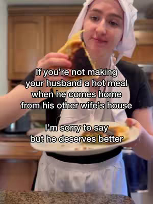A woman wearing a white bonnet and apron holds a plate with a slice of food, possibly a casserole or quiche. She is in a kitchen setting composed of wooden cabinets.