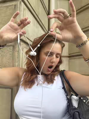A woman with auburn hair and a white top stands in front of a building, making a heart shape with white earphone cords. Her hands and arms are in the frame, adorned with rings and bracelets. The background suggests an outdoor urban setting, with a neutral tones.