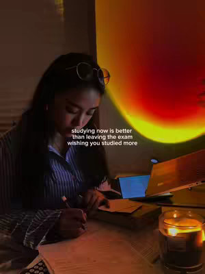A young woman is seated at a desk, illuminated by a warm, orange and yellow light projected onto the wall. She is focused on papers and a laptop, writing with a pen. A candle on the desk adds to the cozy, studious atmosphere.