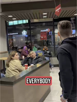 A man standing in front of a group of people seated in a coffee shop. The man is in the foreground, with his back towards the camera. A text overlay 'EVERYBODY' is displayed in a red rectangular box in the bottom center of the image.