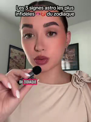 A woman with light skin and dark hair, wearing a light-colored top, is centered in a selfie-style shot. She holds a small microphone close to her lips, and the background includes framed pictures and neutral-colored walls. The image is well-lit, with a focus on her face and expression.