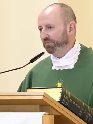 A medium, eye-level shot features a fair-skinned man in a green garment, a white neck collar, and a microphone pointing toward him. He stands behind a wooden podium, partially obscuring a black book labeled "LECTIONARY." The background is a light beige wall.
