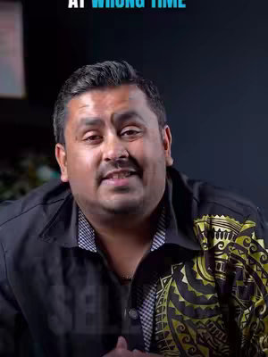 A man with dark hair and a mustache looks toward the camera, set against a blurred background. He is wearing a black shirt, which has a patterned design on the upper right side.