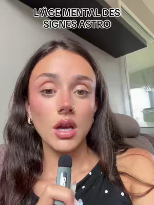 A woman with dark hair and makeup holds a microphone, looking towards the camera in a selfie-style shot. The background shows an interior setting, suggesting a home or office. Overhead text reads: "L'AGE MENTAL DES SIGNES ASTRO".