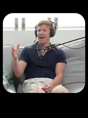 A man with light hair is sitting on a grey couch, wearing a dark blue polo shirt and white shorts. He has headphones on and is speaking into a microphone. In the background is a white wall with some bottles lined up on a shelf. There's a glass jar on the coffee table.