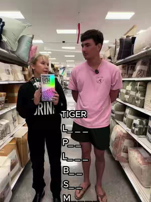 A woman and a man stand in a store aisle, likely a Target, with shelves stocked with pillows and bedding behind them. The woman on the left holds a book with a colorful cover, and a text overlay partially obscures the view of the man, who is in a pink T-shirt and shorts. The text overlay is a fill-in-the-blanks puzzle, probably Tiger - L _ _ - P _ _ - L _ _ - B _ _ - S _ _ - M _ _.