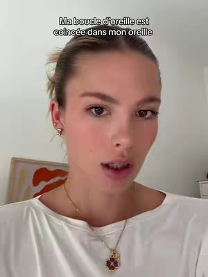 A woman with light skin and flowing brown hair, wearing a white top and a gold necklace with a pendant. Text overlay reads: 'Ma boucle d'oreille est coincée dans mon oreille'. The background has blurred textures. The composition is a close-up, well-lit shot, emphasizing the woman's facial expressions.
