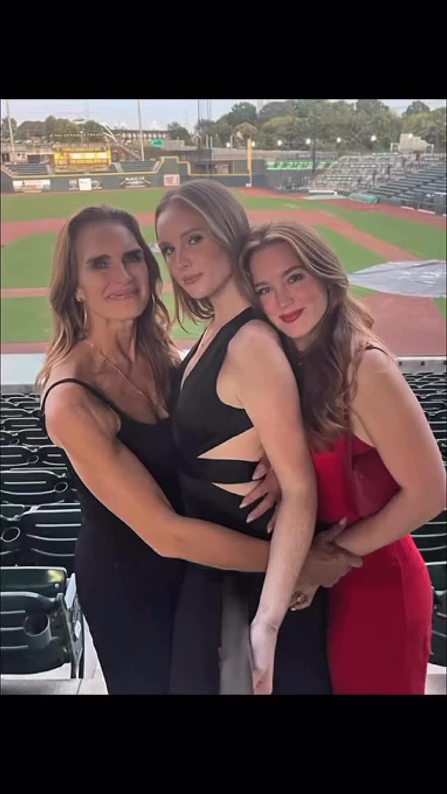 The image shows three women of different ages posing closely together, likely for a photo at what appears to be a sports stadium, with a baseball field visible in the background. They are dressed in what appear to be dresses, one in a black dress with cutouts, the other in a red dress and the last person in a black dress. The women are embracing each other.
