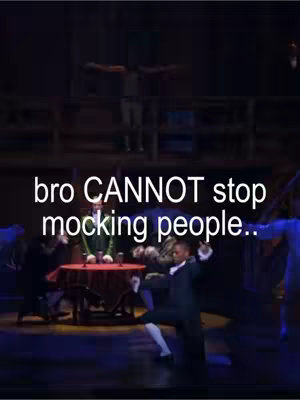 The image captures a low-angle stage shot from a theater production. A group of performers are on stage, with figures in period costumes. In the foreground, a man in a black suit with white accents on his arms is center stage, frozen in an action pose, seemingly dancing. There is text overlayed at the bottom that is white and bolded. The background is blurry, but we can some stage structures there and a person with his arms wide open.