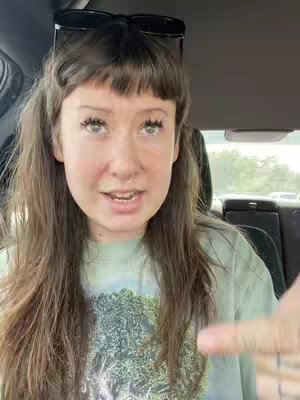 A woman sits in the driver's seat of a car, looking at the camera and talking. She has long brown hair, a nose ring, and is wearing a light green shirt with a tie-dye-like pattern. Sunglasses are perched on her head. The background shows the car's interior, a blurry view of the outside, and parts of the car seat.