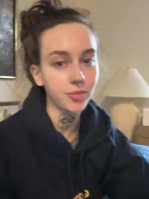 A medium shot shows a woman with dark hair pulled up in a bun, dark-colored hoodie and visible tattoo on her neck. She is looking directly at the camera. The background is a cozy interior with a lamp and framed picture.