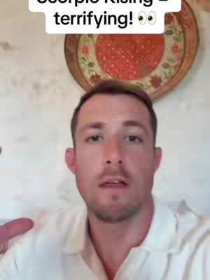 A man is centered in the frame, facing the camera and speaking. Above him, a text overlay reads 'Scorpio Rising - terrifying!'. Behind him, a decorative plate with a red, textured center hangs on the wall. The background suggests an interior setting with off-white walls.