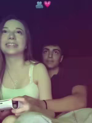 A young woman is holding a video game controller while looking upwards, seemingly engrossed in the game. Behind her, a young man embraces her while looking toward the camera. The background is dark, with a blurred effect.