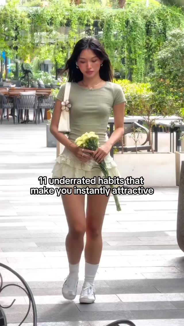 A full-length shot of a young woman walking towards the camera. She holds a bouquet of yellow flowers and is dressed in a sage green top and a white ruffled skirt. The background is blurred but appears to be an outdoor setting with greenery.