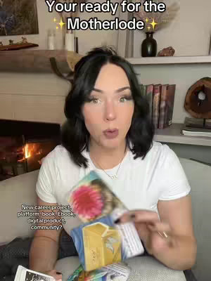 A young woman with dark hair and a white t-shirt is holding a deck of cards, looking directly at the camera with a slightly surprised expression. The backdrop includes a fireplace, bookshelf, and various decorative items.