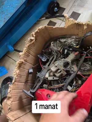 A first-person perspective view inside a cardboard box filled with various motorcycle parts and wires. A hand, presumably the creator's, touches a red plastic component. A blue object with wheels is visible in the background. A text overlay stating "1 manat" appears in a white rectangular box.