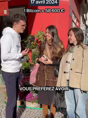 A man in a white hoodie interviews two women in front of a red-walled building with a flower shop. On the top left, text indicates the date and bitcoin's value. The lower part of the image has text. The man holds a microphone.