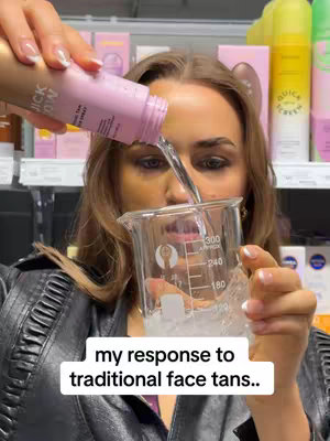 A woman is pouring a clear liquid from a pink and gold spray bottle into a clear beaker. The woman is in a store setting with shelves in the background. Text overlayed across the bottom reads 'my response to traditional face tans..'.