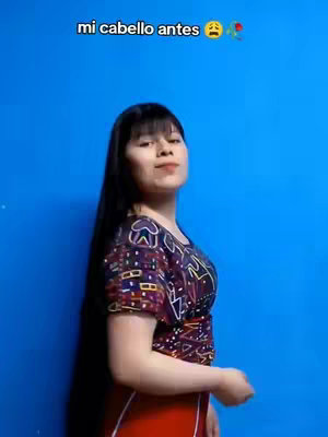 A woman with long, dark hair and bangs poses in front of a bright, blue background. She wears a colorful top and red skirt, her expression neutral. Text overlayed on top reads, "mi cabello antes".