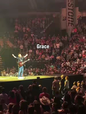 A wide shot of a man on a stage with arms outstretched, playing a guitar in front of a large crowd. The word "Grace" is overlaid on the image.