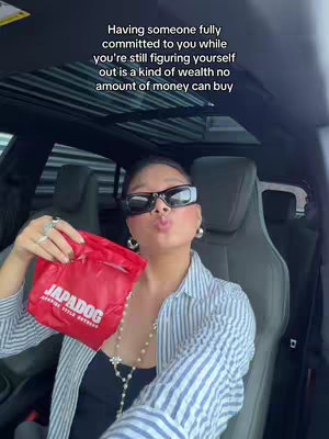 A person is seated inside a car, facing the camera with a pout. They are wearing sunglasses, a striped shirt, and holding a red bag. Text is overlaid at the top.