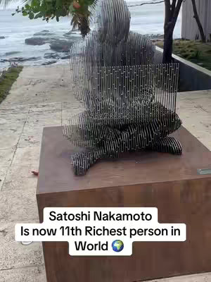 A shot of a metal sculpture of a figure seated on a pedestal. The sculpture appears to be made of thin, vertical metal strips, giving it a semi-transparent appearance. The sculpture is positioned in front of a body of water with a sky that suggests a sunset or sunrise, some trees in the background. A text overlay is present, with the words 'Satoshi Nakamoto Is now 11th Richest person in World' written in a white font.