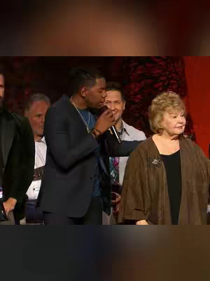 A group of individuals on stage, including a Black man with a microphone, a woman in a brown jacket, and other men. The setting appears to be a performance or event backdrop with red and dark elements, with individuals in formal attire.