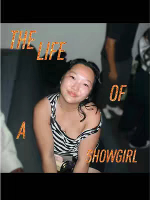 The image captures a young woman seated with a casual, candid vibe. She's positioned against a blurred background, possibly indoors, with indistinct figures partially visible. The woman is wearing a striped tank top. Overlaying the image is the text 'THE LIFE OF A SHOWGIRL' in a glittering, orange font, the word 'OF' is placed vertically.