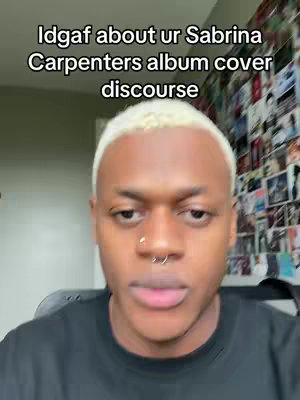 A close-up shot of a young Black man with short, bleached blonde hair and a nose ring. He is looking directly at the camera with a neutral expression. Behind him, a wall is covered with what appears to be various photos and images. The overall composition is simple, focusing on the man's face and conveying a sense of direct communication.