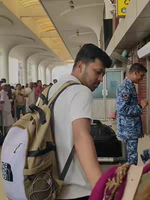 A man with dark hair and a backpack walks through a brightly lit airport concourse. He is wearing a white t-shirt. People are walking in the background.