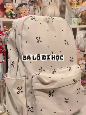 The image is a close-up of a white backpack with a pattern of small black bows and dots. The backpack has a large main compartment and a front pocket. The text "BA LÔ DI HỌC" is overlaid on the backpack in a rectangle.