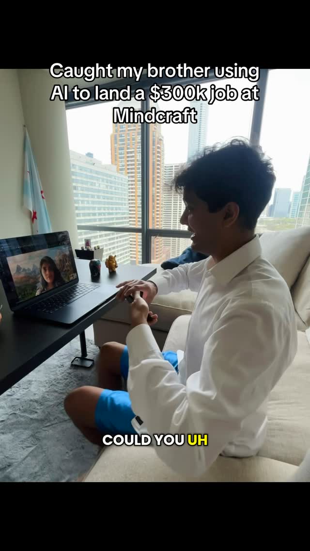 The video shows a man sitting in a well-lit room, looking towards a laptop screen. The man is wearing a white shirt and blue shorts. The laptop displays a video call with a woman on the screen. The room has a large window offering a city view in the background and a flag hanging on the wall. A desk is in front of the window with some decor on top of the table.
