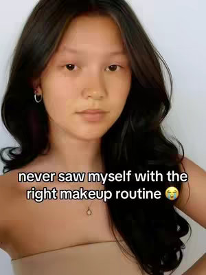 A young woman with long, dark hair and fair skin looks directly at the camera. Text overlayed reads, "never saw myself with the right makeup routine" above her. She wears a light-colored strapless top and a delicate necklace. The background is a plain, light color.