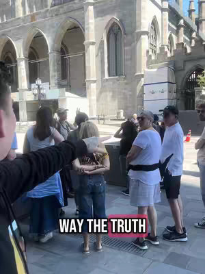 A person with their arm extended is pointing at the words "WAY THE TRUTH" overlaid on the image. The foreground features a person with the arm. In the background, a group of people stands in front of and beside a stone building on a bright day.