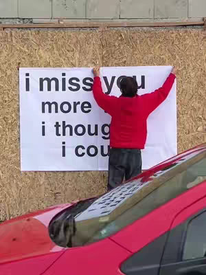A person in a red hoodie is standing in front of a plywood wall, holding up a white banner with black text that states 'i miss you more i though i cou'. A red car is partially visible in the foreground.