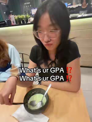 A young woman with long black hair and glasses is seated at a table, possibly in a restaurant, looking down and to the right. She is holding a spoon over a black bowl containing a green, creamy substance. The background shows a beige wall and other customers, with overhead lighting.