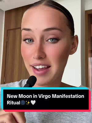 A woman with fair skin and light-colored eyes is looking directly at the camera, smiling slightly and speaking. The background is a neutral tone; the woman is wearing a grey top. Below her face is a rectangular, black, semi-transparent box with white text. The text says, "New Moon in Virgo Manifestation Ritual." Below this is a row of graphics: a crescent moon, some star bursts, and a white heart icon at the end.