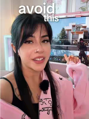 A woman with dark hair and a nose ring is talking while pointing towards the screen. The hook 'avoid this' is placed above her head in white font. The background shows the woman's room, which contains a computer screen showing a game and a pink setup. The woman is wearing a black top and a pink zipped jacket.