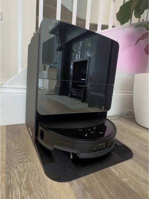 A black robot vacuum cleaner and its cleaning/charging station are displayed on a light wooden floor. The vacuum sits on a black base, with a black, glossy upper section that appears to house its controls. In the reflection of the gloss black surface, the interior of a room is visible. The background includes a white staircase with wooden steps, the right part of the scene is occupied by a white pot and lush green plant.