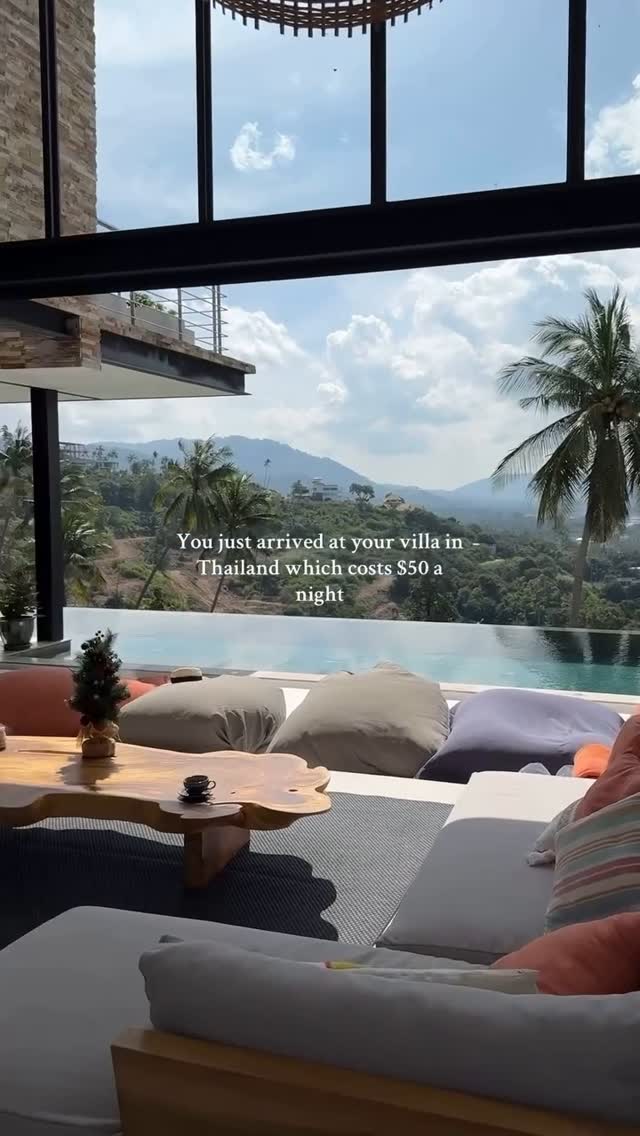 An outdoor shot taken from a covered patio looking out towards a scenic vista. A zero-edge pool is in the foreground. Light-colored, oversized bean bags are set out on the patio with a wooden table and coffee cup. Beyond the pool, palm trees, lush greenery, and rolling hills are visible under a lightly clouded sky. The composition features a sense of luxury and relaxation.